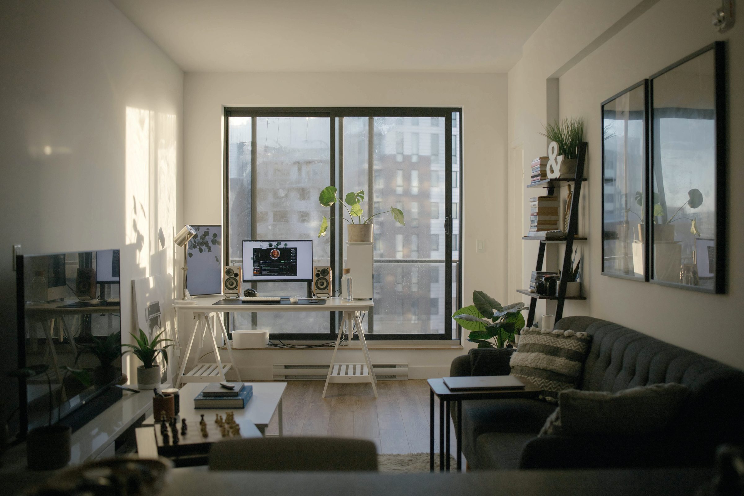 A sunlit apartment with a desk at the window, plants and a low sofa.