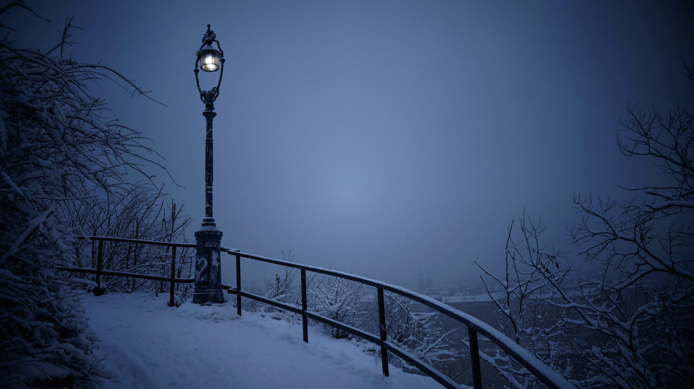 A lamplit path above Prague, railings and branches under fresh snow.
